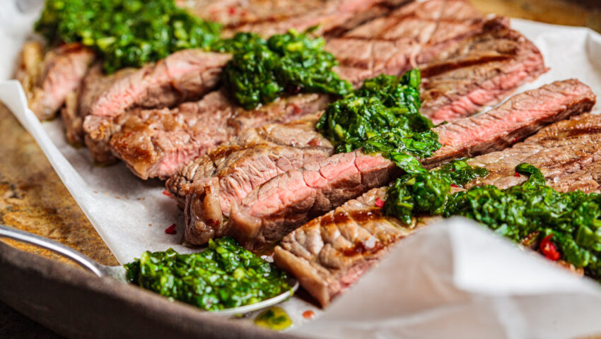 Sliced grilled beef steak with chimichurri sauce on a dark dish, dark background.